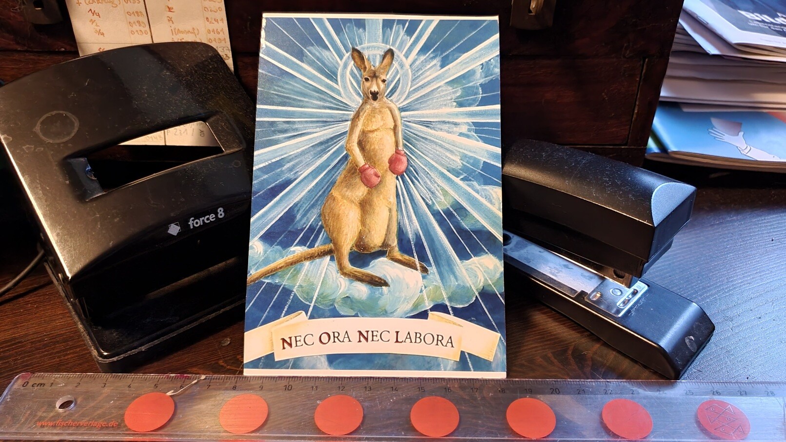 Arrangement mit Locher, Tacker, Lineal und einer Postkarte vom Känguru mit Heiligenschein und dem Slogan 'nec ora nec labora'