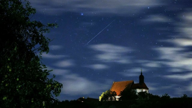 Horizontbild mit einer etwas entfernt stehenden beleuchteten Kirche. Darüber ein Nachthimmel mit einzelnen Zirruswolken. Die Leuchtspur einer Sternschnuppe und einzelne Sterne sind erkennbar.