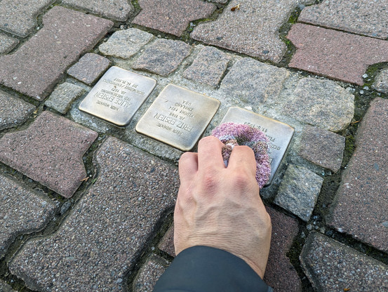 Die linke Hand von Daniel R. Schmidt wischt mit einem alten Lappen über drei in einen Bürgersteig eingelassenen Stolpersteine. Die Inschrift steht auf dem Kopf. Die Stolpersteine sind für Familie Epstein verlegt worden, wie die Inschrift erahnen lässt. Die Steine leuchten frisch gereinigt und golden.