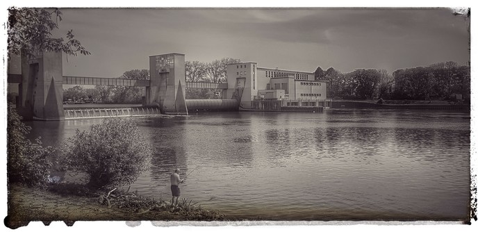 Schwarz-weiß Foto mit Blick über den Main auf die Staustufe in Eddersheim. Im Vordergrund am Ufer ein Angler.