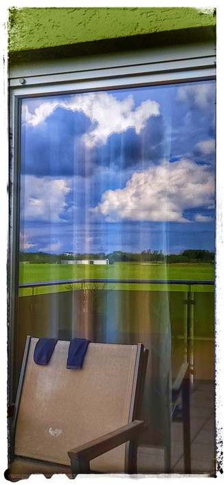Foto eines Balkonfensters, vor dem ein Stuhl steht, auf dessen Lehne ein Paar Socken lüftet. Im Fenster spiegeln sich die Felder hinter dem Haus, über denen der blaue Himmel mit weißen und grauen Wolkenbänken erstrahlt. Zwischen den Feldern leuchtet ein landwirtschaftlicher Geräteschuppen in weiß.