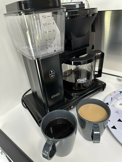 Eine schwarze Moccamaster mit frisch durchgelaufenem Kaffee, davor zwei graue Tassen aus Kunststoff, links Kaffee pur, rechts Milchkaffee. 