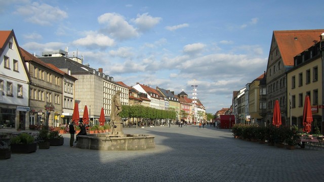Das Bild zeigt die Maximilianstraße in der gesuchten Stadt 2014, vorne ein Brunnen in der Fußgängerzone, weiter hinten steht ein Maibaum.