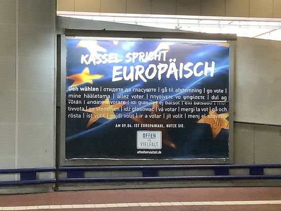 Plakat vor dem Hintergrund des Stoffs einer EU Flagge steht 

Kassel spricht europäisch
„Geh Wählen“ in allen Sprachen und Alphabeten der EU , getrennt durch senkrechte Striche Am 9. Juni 2024 ist Europawahl. Nutze sie
Weißes Feld
Offen Für Vielfalt

OffenFuerVielfalt.de
