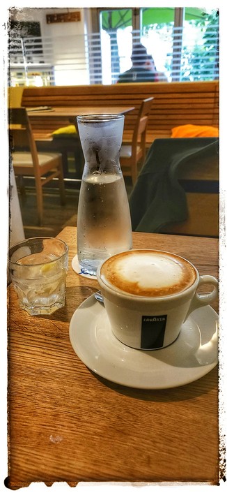 Tasse Milchkaffee und eine Karaffe Wasser, nebst Glas auf einem Tisch. Im Hintergrund Tische und Stühle im Café. 