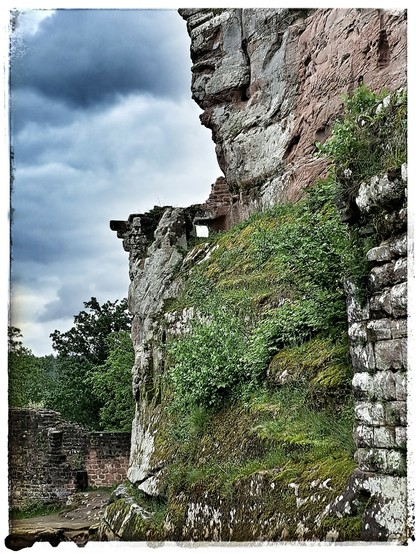 Teil der Ruine Burg Fleckenstein. Eine fast quadratische Fensteröffnung in einer verfallenen Mauer, fast in der Mitte des Fotos. Unterhalb sind rechts Mauern und Felsen zu erkennen, die teilweise von Pflanzen, Flechten und Moosen bewachsen sind; unten links ist im Hintergrund ein teil einer Burgmauer etwas jüngeren Datums zu erkennen. Oberhalb rechts ist der rot-graue Sandstein zu erkennen, in den die Felsenburg gegraben wurde. Der Himmel ist dunkler, dramatischer per Bildbearbeitung etwas verf…