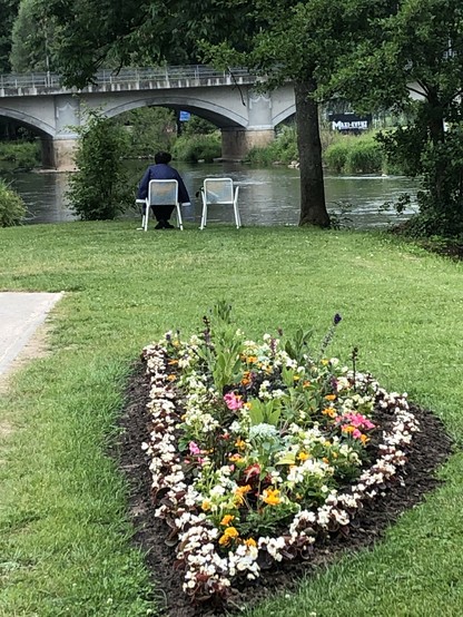 Blick über ein Blumenbeet über Wiese auf einen Fluss im Hintergrund eine Brücke. Am Ende der Wiese stehen zwei weiße Gartenstühle, darauf sitzt links eine Frau. Von hinten.
