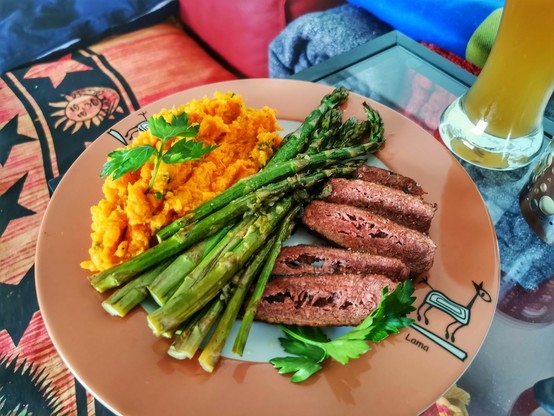 Plant based steak, aufgeschnitten, gegrillte grüner Spargel und Süßkartoffelpüree, angerichtet auf einem Teller mit ockerfarbigem Rand und einem stilisierten Lama