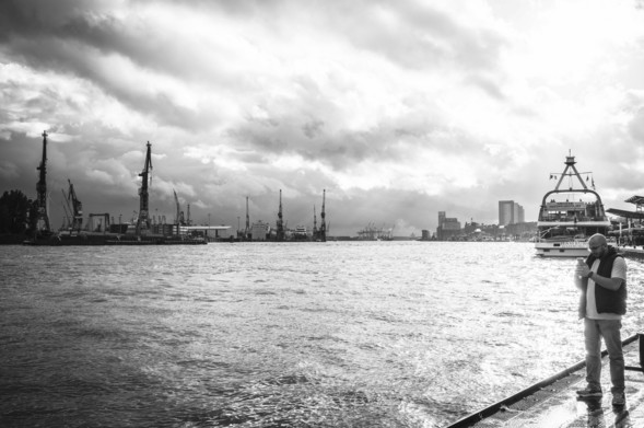 Eine Person steht am Ufer der Elbe (Landungsbrücken) in Hamburg. Ein dramatischer Himmel, von der untergehenden Sonne in Szene gesetzt - das Foto in schwarz-weiß.