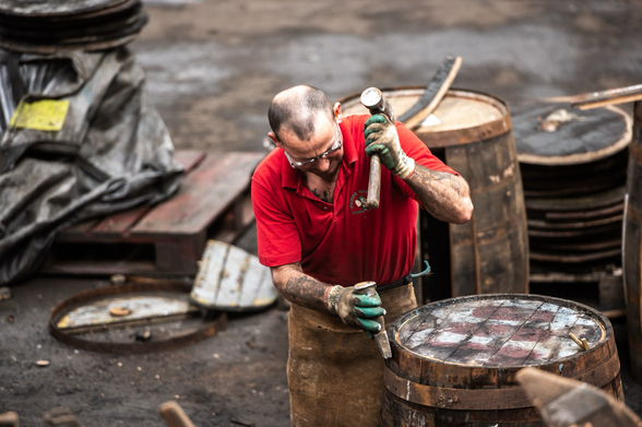 Im Vordergrund dieses Fotos ist ein Mann mit Lederschürze und roten Poloshirt zu sehen. Dieser Mann bearbeitet mit Werkzeug ein vor ihm stehendes Whiskyfass.