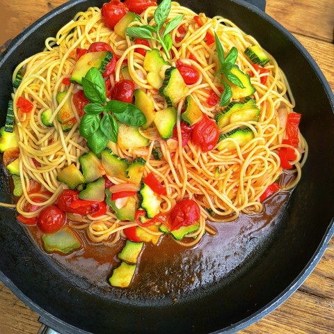 In einer schwarzen Pfanne liegen Spaghetti mit gebratenen Zucchini, Zwiebeln, Paprika, Tomaten. 