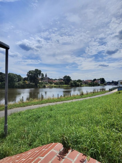 Blick über die Uferlandschaft der Weser. Alles grün und kleiner Radweg. Am anderen Ufer Hoya mit Kirchturm.