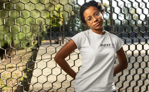 Eine schwarze, weiblich gelesene Person vor einem Gitter im Freien. Auf ihrem T-Shirt steht der Schriftzug 