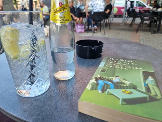 Man sieht ein Buch von Max Gold und einen Gin Tonic auf einem Außentisch in einer Bar.
Dahinter großstädtisches Leben.