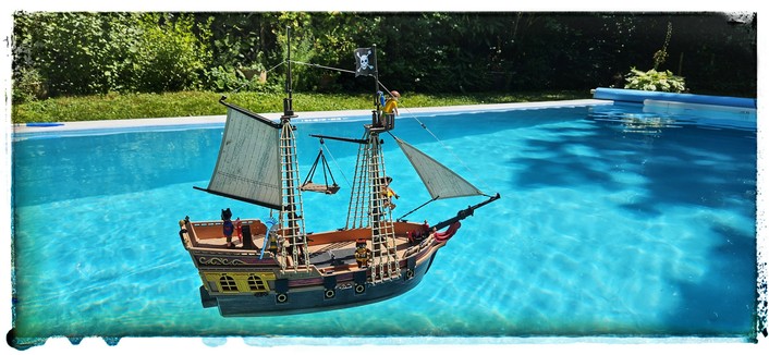 Piratenschiff aus dem Hause Playmobil auf dem Schwimmbad im Garten. Die Crew besteht aus diversen Figuren aus anderen Sets, u.a. ein Bauarbeiter mit blauem Blätterrechen (Kapitän Strabag), ein Mädchen in Sommerklamotten und diverse Piraten.
