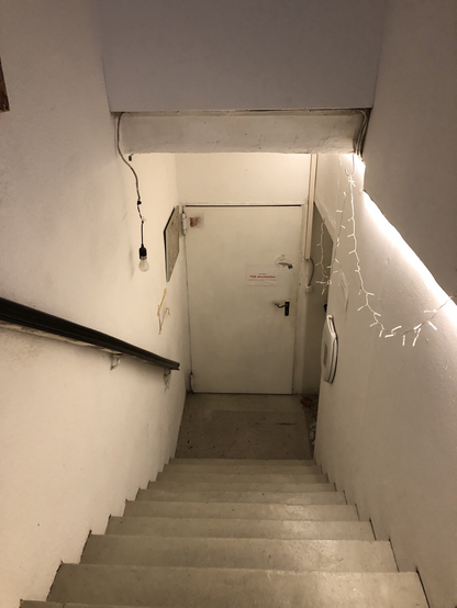 Treppe von oben links ein Geländer rechts weiße Wand und einen Klodeckel (dahinter verbergen sich die Kloschüssel) und eine Lichterkette unten eine Metalltür