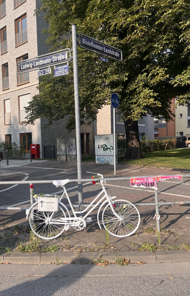 Ein mit Blumen dekoriertes weiß lackiertes Fahrrad steht angelehnt an ein Gehweggeländer. am Rad ist ein Schild angebracht, auf dem steht: Radfahrer, 41 Jahre, 09.06.2024. Hinter dem Rad zeigt ein Straßenschild die Straßennamen „Ludwig-Landmann-Straße“ und „Rödelheimer Landstraße“