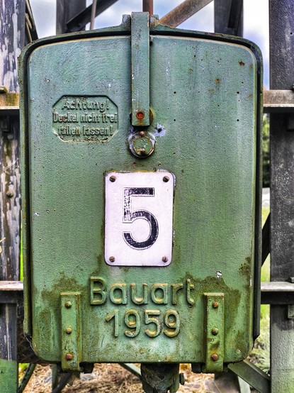 Ein grün lackierter Schaltkasten der Firma AEG hängt an einem Fagrdrahtmast der Bahn. Auf seinem Deckel eingeprägt steht 