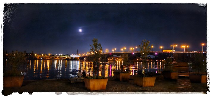 Blick auf die nächtliche erleuchtete Theodor Heuss Brücke. Die Lichter der Brücke und die Beleuchtung am Ufer in Kastell spiegeln sich im Wasser. Im Vordergrund stehen kleine Bäume in Kübeln. Über der Szene steht der Mond.