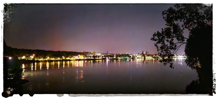 Blick von der Eisenbahnbrücke über den Rhein auf das nächtliche Mainz. Die Gebäude entlang des Ufers sind hell erleuchtet und das Licht spiegelt sich im Fluss. Der Himmel wirkt lila mir vereinzelten Sternen. Die Wasseroberfläche ist spiegelglatt und ruhig. Rechts im Bild ein Baum.