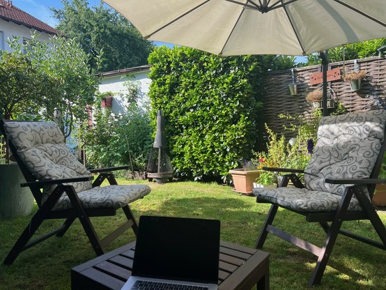 Blick in einen Reihenhausgarten mit Rasen, Hecken, Sonnenschirm und Gartenstühlen. Im Vordergrund ein Laptop auf einem Gartentisch.