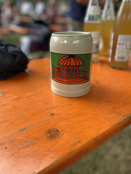 Ein keramischer Bierkrug mit einem bunten Logo sitzt auf einem orangefarbenen Holztisch. Im Hintergrund gibt es verschwommene Bilder von anderen Getränken und Menschen.