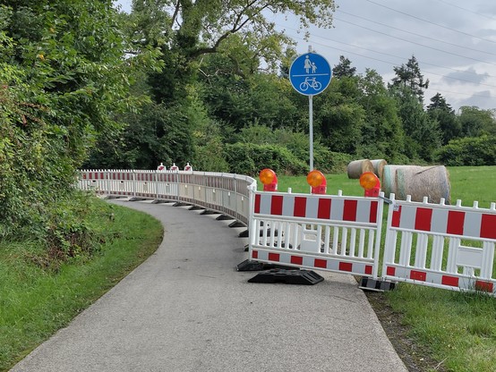 Feldweg im Grünen, der durch rot-weiße Kunststoffelemente halbseitig gesperrt ist.