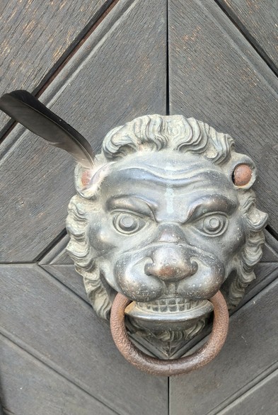 Ein Löwenkopf an einer Tür mit Türklopferring im Maul. An seinem linken Ohr steckt eine dunkle Feder.