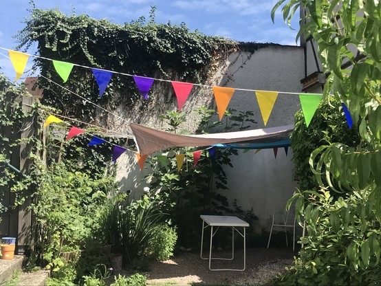 Ein sonniger Innenhof mit etlichen Pflanzen, überspannt von Sonnensegeln und bunten Wimpeln. Auf einem Kiesboden ein weißer Tisch, im Hintergrund ein weißer Gartenstuhl.