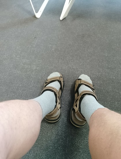 Zwei Beine mit grauen Socken und Sandalen auf einem grauen Teppichboden 