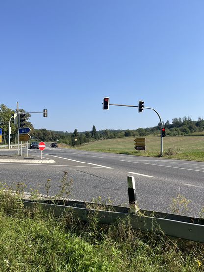 Bundesstraße ohne Radweg mit Wegweisern wird von zwei Autos befahren. Rechts eine rote Ampel. Im Hintergrund Wald und eine frisch gemähte Wiese