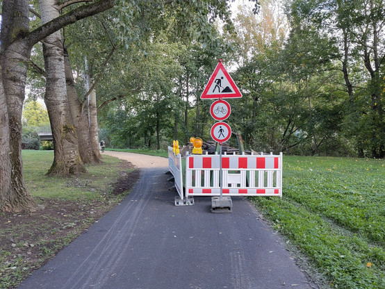 Asphaltierter Weg in einem Park, der an einer Baustellenabsperrung endet. Dahinter wird der Weg mit sogenannter 