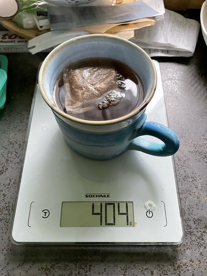 A blue ceramic mug filled with tea is sitting on a digital scale displaying 404 grams. A tea bag is submerged in the tea, with bubbles rising to the surface. Various kitchen items are visible in the background.