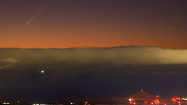 Der Komet ist relativ klein dicht am Horizont am rötlich gefärbten Himmel sichtbar. Über der Landschaft im Bildhintergrund liegen Wolken oder Nebel. Am unteren Bildrand sind Lichter einer Ortschaft zu sehen