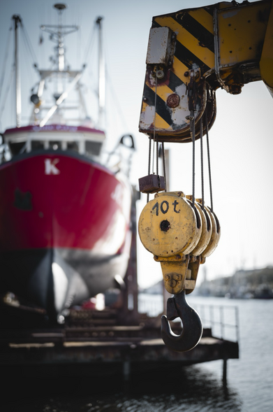 Im Vordergrund hängt ein gelber Haken von einem Kran herab. Er trägt die Aufschrift 10 t. Im Hintergrund sieht man ein rotes Schiff.