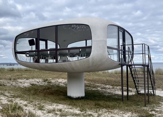 Farbiges Querformat. Ein Bauwerk mit großen Fenstern und einer Metalltreppe rechts steht auf einer Düne. Dahinter ein schmaler Streifen Meer, darüber Himmel und Wolken. Das Bauwerk ruht auf einem zentralen Pfeiler, besteht nur aus einem einzigen Raum mit Fenstern nach allen Seiten und ist vom Dach, Boden und Ecken her abgerundet. 