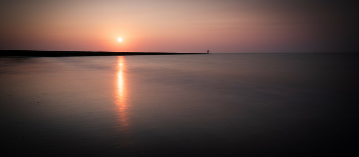 Die Sonne geht über dem Meer unter, das sich flach in Richtung Horizont ausbreitet. In der Ferne sieht man eine Buhne, die sich ins Meer erstreckt und an deren Ende ein Mann angelt.