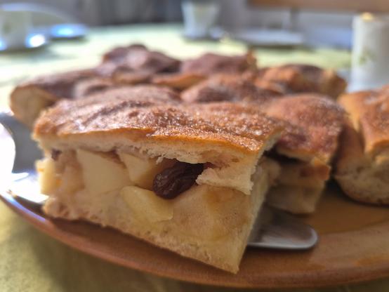 Mehrere Stücke gedeckte Apfelkuchen auf einem brauen Teller. Das vordere Stück im Fokus liegt auf einem silbernen Kuchenheber. Der Hefeteig des Deckels ist goldbraun. In der Füllung des vorderen Stücks ist auch eine Rosine zu erkennen. Die Apfelstücke sind ca. 2 cm groß. 