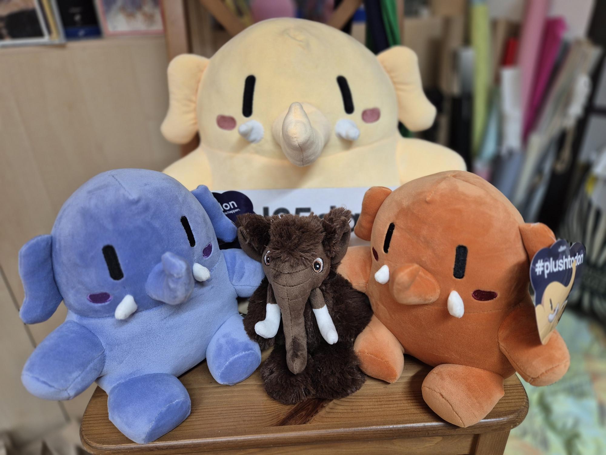 Auf einem Stuhl sitzen ein großes und zwei kleinen Plushtodons. Dazwischen ein anderes Plüsch-Mammut.