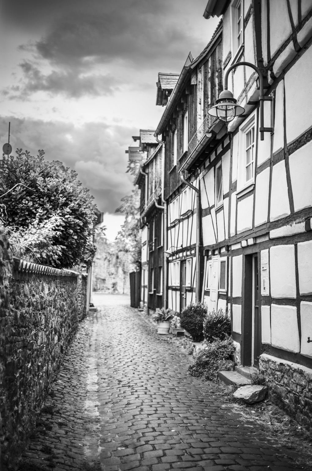 Das Foto zeigt eine schmale, kopfsteingepflasterte Gasse zwischen alten Fachwerkhäusern. Die Aufnahme ist in Schwarz-Weiß, was der Szene einen zeitlosen, fast historischen Eindruck gibt.  Rechts stehen dicht an dicht mehrere Fachwerkhäuser. Ihre weißen Wände sind mit dunklen Holzbalken durchzogen, die unregelmäßige Muster bilden. Viele kleine Fenster blicken zur Gasse, und an einer Hauswand hängt eine schlichte, runde Laterne aus Metall und Glas.  Links verläuft eine alte Steinmauer, deren Oberfläche uneben und rau ist. Dahinter ragen Büsche und Blätter hervor. Die Gasse wirkt eng, aber gepflegt, und sie führt leicht geschwungen in die Tiefe des Bildes, wo Bäume und weiteres Mauerwerk zu erkennen sind.  Der Himmel ist wolkig und wirkt schwer, als würde gleich ein Gewitter aufziehen. Durch die Schwarz-Weiß-Wiedergabe wirken die Wolken dramatisch und kontrastreich.  Insgesamt vermittelt das Foto eine ruhige, fast stille Stimmung – wie ein Moment in einem kleinen, historischen Dorf, früh am Morgen oder kurz vor einem Wetterumschwung.
