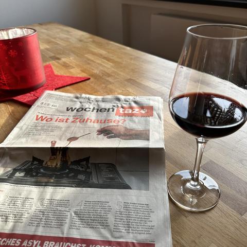 Auf einem hellen Holztisch liegt eine Ausgabe der Wochenzeitung „wochen taz“ mit der Schlagzeile „Wo ist Zuhause?“ vom 20.–26. Dezember 2025. Daneben steht ein Glas mit Rotwein, das etwa zur Hälfte gefüllt ist. Links im Bild sieht man eine rote, dekorative Kerzenhalterung mit einem kleinen Stern auf einem passenden Untersetzer. Die Atmosphäre wirkt gemütlich und einladend.