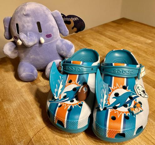 Auf dem Bild sitzt ein niedliches , lila Stofftier (Elefant) mit großen Augen und kleinen Stoßzähnen auf dem Tisch. Daneben befindet sich ein Paar bunte Crocs-Schuhe, die mit dem Logo und den Farben der Miami Dolphins (ein NFL-Football-Team) verziert sind. Die Schuhe sind weiß mit orange-blauen Streifen und haben mehrere Anstecker, darunter ein Mini-Football und ein Logo der Dolphins.