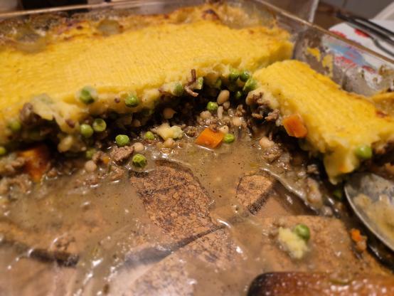 Shepherd's Pie in einer Auflaufform aus Glas. Nur noch wenige Stücke sind übrig. Goldfarbene Kartoffelbreidecke auf Mischung aus Hackfleisch, Karotten, weißen Bohnen, Zwiebeln, Erbsen, Rosmarin. Rechts im Bild der vordere Teil eines Esslöffels und der vordere Teil eines Holzschabers, der vorne von der häufigen Benutzung dunkler verfärbt ist. Zwei Korkuntersetzer scheinen durch den Boden der Form durch. Die Holz-Tischplatte darunter ist braun. Rechts ist ein weißer Teller mit rot-hellblauer flor…
