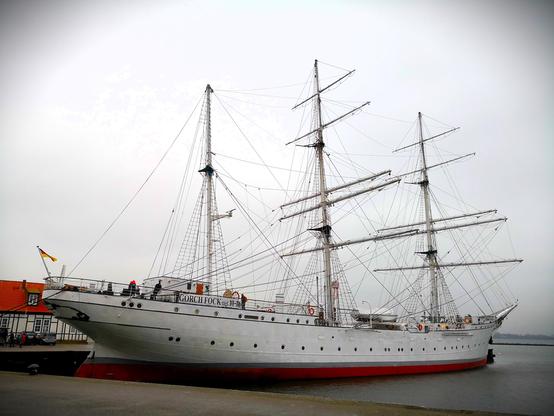 Farbiges Querformat: Dreimastiges Segelschulschiff Gorch Fock im Hafen, mit deutscher Flagge am Heck. Der Rumpf ist weiß, knapp über der Wasserlinie verlaufen ein grauer und roter Streifen. Auf dem Foto sind keine Segel gesetzt, das Schiff liegt im Hafen. Links ein Fachwerkhaus mit rotem Dach.