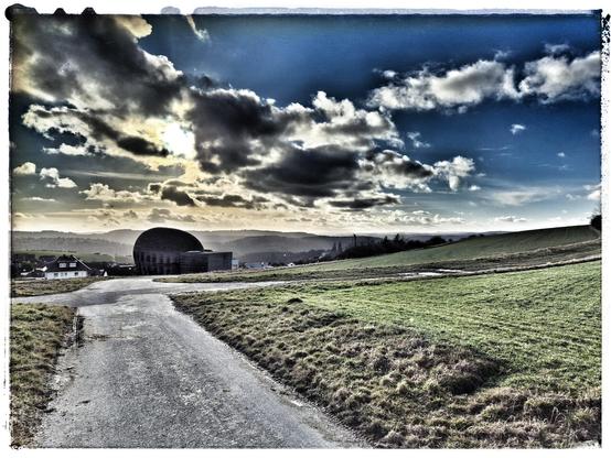 Feldweg und Felder auf der Höhe. Eiförmig Kapelle aus Holz als schwarze Silhouette. Hinter der Kapelle Wohnhäuser in Langenseifen. Am Horizont Mittelgebirgskette (Hunsrück). Leichte Hochnebel in den Tälern im Hintergrund sorgen für eine diffuse Beleuchtung. Einzelne Wolken und blauer Himmel. Die Farben sind dramatisch dunkel verfremdet. Sonne bricht durch die dunklen Wolken. 