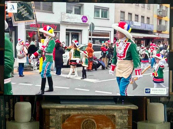 Ein buntes Bild eines Fastnachtsumzugs in der Stadt: Eine Gruppe fröhlich kostümierter Teilnehmer:innen marschiert in prächtigen Uniformen mit hohen Hüten, Federn und farbenfrohen Jacken durch die Straßen. Im Hintergrund sind Zuschauer:innen zu sehen, die den Umzug begeistert verfolgen, während Geschäfte wie "Asia Family" im Bild zu erkennen sind.