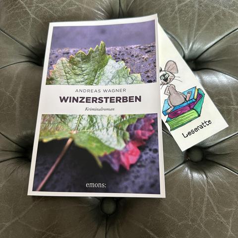 Die Softcoverausgabe von ANDREAS WAGNERs Kriminalroman WINZERSTERBEN aus dem emons: Verlag liegt auf einem kapitonierten Hocker mit grünem Leder, darin ein handgemaltes Lesezeichen mit einer bunten Leseratte auf einem Bücherstapel. 