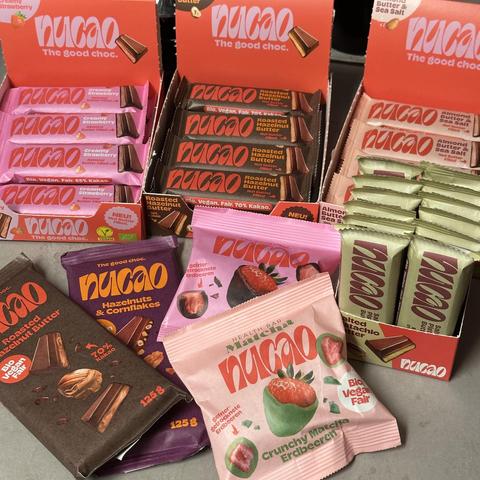 Das Bild zeigt eine bunte Auswahl an veganen Schokoriegeln und -tafeln der Marke **nucao** in verschiedenen Geschmacksrichtungen:

- **Creamy Strawberry** und **Almond Butter Filled** (rosa Verpackung)
- **Roasted Hazelnut Butter** (dunkelbraune Verpackung)
- **Hazelnuts & Cornflakes** (lila Verpackung)
- **Crunchy Matcha Erdbeeren** (pink-grüne Verpackung)
- **Salted Almond Butter & Sea Salt** (hellgrüne Verpackung)

Die Produkte sind bio, vegan und fair gehandelt, mit einem Kakaoanteil von bi…