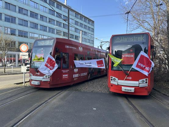 "Zwei rote Straßenbahnen stehen auf Gleisen in einer Stadt und nehmen an einer Streikaktion teil. Beide Straßenbahnen tragen Schilder mit der Aufschrift 'Heute STREIK' und 'Warnstreik'. An den Fahrzeugen sind Fahnen der Gewerkschaft ver.di befestigt, eine davon mit der Aufschrift 'Dieser Betrieb wird bestreikt'. Im Hintergrund sind Gebäude und ein Verkehrsschild zu sehen."