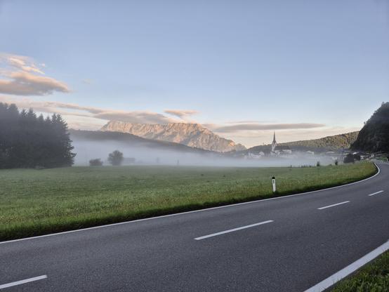 Morgenstimmung mit Blick auf Adnet und den Untersberg. Nebel liegt im Tal über sattgrünen Wiesen. Im Vordergrund eine zweispurige Landstraße. Der Untersberg wir von der Morgensonne beleuchtet. Von dem Ort Adnet ragt ein Kirchturm aus dem Nebel und Häuser sind zu erkennen. Links im Bild ein kleines Tannenwäldchen.  Der Himmel ist hellblau, nur wenige flache Schönwetterwolken. 